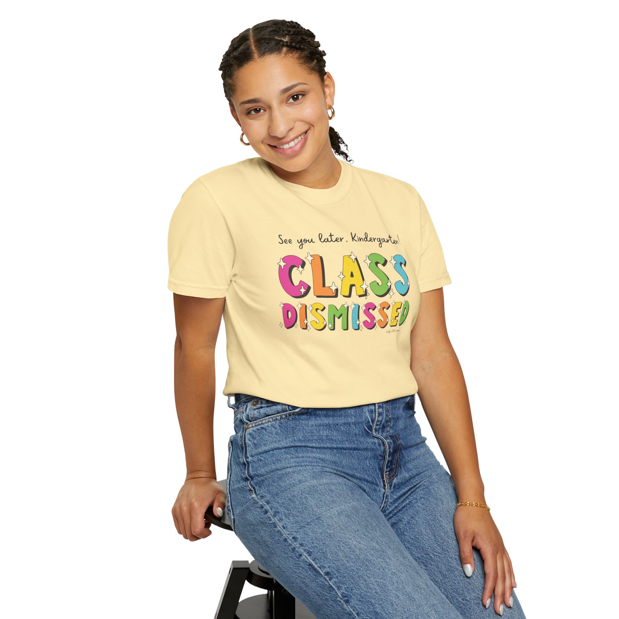 A cheerful young girl wearing a yellow "Class Dismissed" kindergarten T-shirt, smiling and sitting on a stool, showcasing fun and colorful educational apparel for early learners.