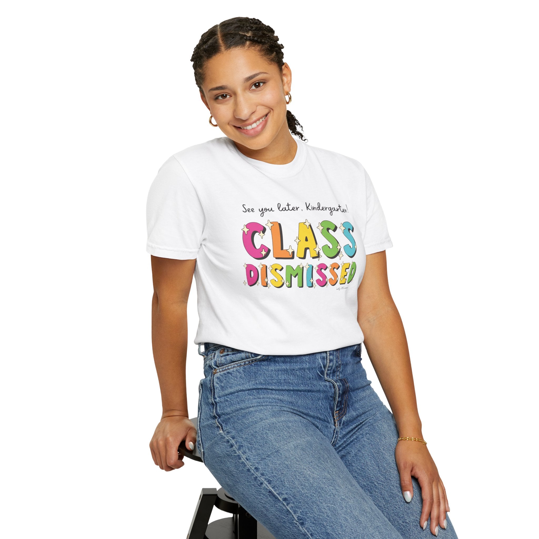 A colorful kindergarten T-shirt with "Class Dismissed" text, worn by a smiling girl sitting on a stool, perfect for celebrating the end of the school year and suitable for preschool and elementary st.